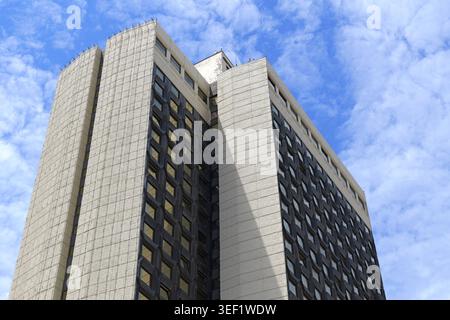 Moderne Hochhausfassade aus Beton und Glas, die sich nach oben hin zu einem klaren blauen Himmel mit weißen Wolken erstreckt und die städtische Entwicklung und Unternehmensgründung symbolisiert Stockfoto