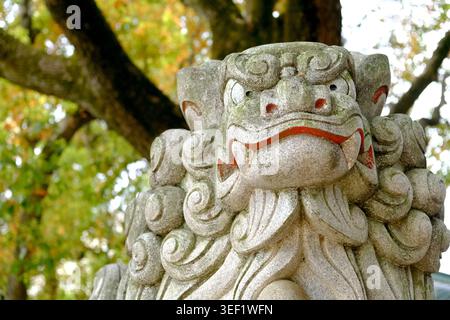 Nahaufnahme einer traditionellen Komainu-Schutzlöwen-Statue aus Stein an einem japanischen Schrein mit Bäumen im Hintergrund Stockfoto