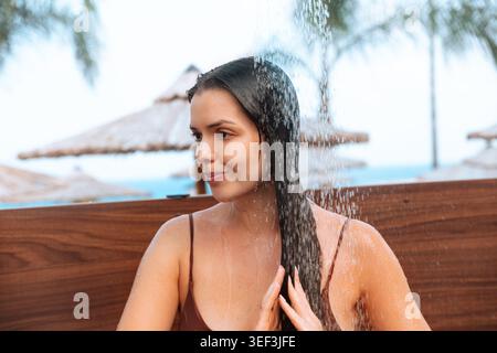 Junge Frau glättet langes nasses Haar unter Sonnenlicht. Attraktive Frau, die sich neben einer tropischen Dusche mit frischem Wasser erholt. Beauty-Lifestyle-Konzept. Stockfoto