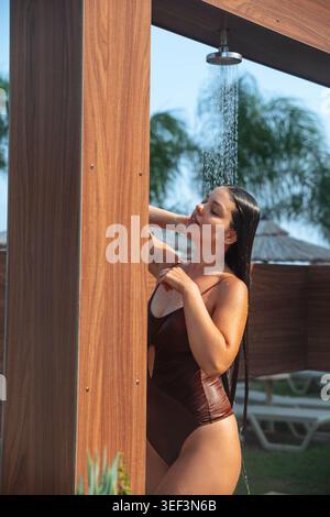 Junge Frau, die in der Nähe einer tropischen Badedusche in stilvollem nassem Badeanzug steht. Attraktive Frau, die sich im Freien mit strahlendem Teint und brünetten Haaren entspannt Stockfoto