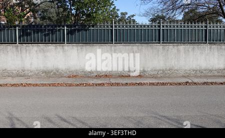 Eleganter Hauszaun mit Metallgeländer und schwarzer Platte an Betonwand. Bürgersteig aus Zement mit heruntergefallenen Blättern und Straße davor. Bäume und Himmel Stockfoto