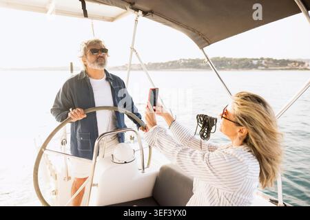 Reifes Paar segelt im Ozean, Frau filmt Mann, der das Boot steuert Stockfoto