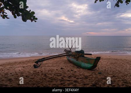 Traditionelles Auslegerboot parkt am Sandstrand von Tangalle bei Sonnenuntergang unter bewölktem Himmel in der Südprovinz Sri Lanka Stockfoto