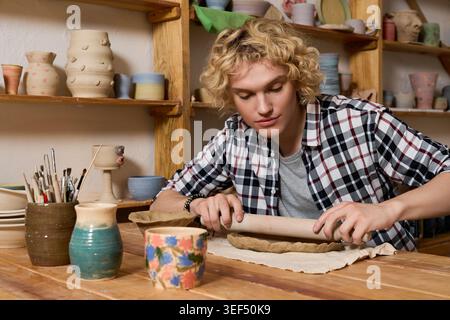 Lockige blonde Töpfer arbeitet intensiv daran, Ton in einer gemütlichen und farbenfrohen Werkstatt zu Formen. Stockfoto