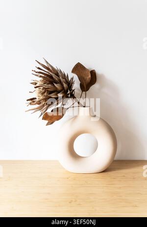 Weiße runde Keramikvase mit getrockneten Protea-Blüten mit Blättern auf neutralem Hintergrund, minimalistisches Stillleben mit weichen Schatten und natürlichem Dekor. Vor Stockfoto