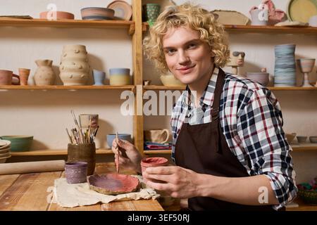 Ein junger Mann formt Ton in einer lebendigen Töpferwerkstatt voller Kreationen. Stockfoto