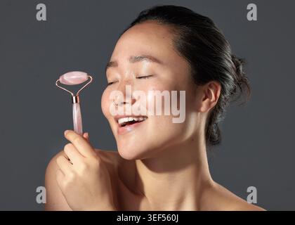 Frau, Derma Roller und Hautpflege mit Lächeln für Durchblutung, Anti-Aging oder Gesichtsheilung im Studio. Stein, asiatische Person und Rosenquarz mit Stockfoto