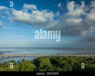 Der Verkehr bewegt sich entlang des Corredor sur Highway über den Ozean in Panama City, mit üppigen grünen Bäumen im Vordergrund und einem riesigen Himmel über dem Meer. Stockfoto