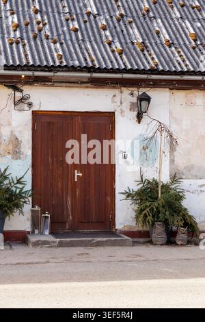 Traditionelle dunkelbraune Holztüren eines verwitterten weißen Bauernhauses mit rustikalen Dekorationen und Vintage-Laternen an einem sonnigen Tag. Stockfoto