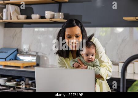 Afroamerikanische Mutter, die eine kleine Tochter hält und auf einen Laptop in der Küchenzeile in der Nähe des Kochfeldes blickt Stockfoto