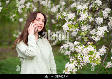 Lächelnde junge Bloggerin, die Videoanrufe macht oder Selfie auf dem Mobiltelefon macht, im Frühlingsgarten in der Nähe von weißen Blütenbäumen Stockfoto