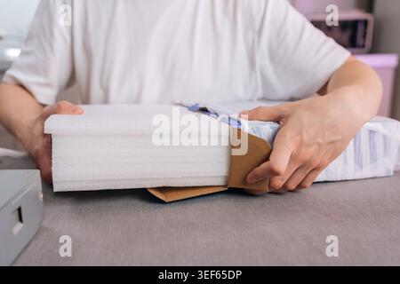 Nahaufnahme der Hände, die Neukäufe aus dem Versandkarton auspacken. Weißer Schutzschaum sichert den Inhalt. Konzept für E-Commerce, Versand, Konsum Stockfoto