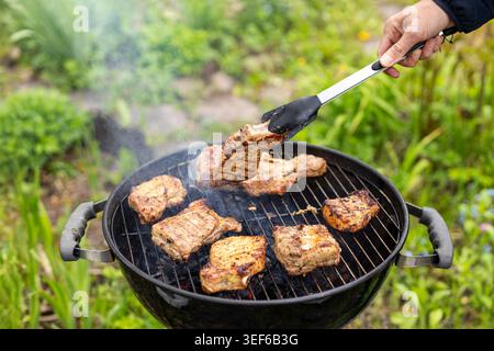 Gegrilltes Fleisch kochen auf dem Grill. Nahaufnahme von rauchigem gegrilltem Fleisch, das mit einer Zange auf einem Holzkohlegrill in einem grünen Garten gedreht wird. Stockfoto
