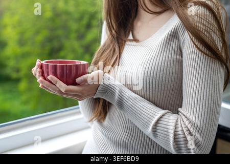 Frau hält warmen Tee am Fenster. Nahaufnahme einer Frau in einem gemütlichen Pullover mit einer roten Tasse in der Nähe eines Fensters mit grüner Natur draußen. Stockfoto