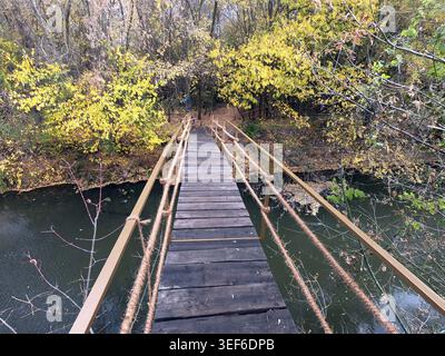 Schmaler Fußgängersteg mit Holzdielen und Seilgeländern, der einen dunklen Fluss überspannt, umgeben von Bäumen mit gelbem und grünem Herbstlaub in einem friedlichen n Stockfoto