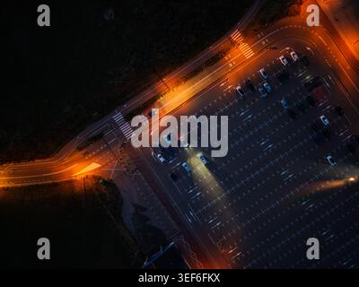 Dramatischer Blick von oben nach unten auf einen Parkplatz bei Nacht mit leuchtenden Straßenlaternen. Die Autoscheinwerfer beleuchten den Asphalt in der Nähe einer Straßenkreuzung (cre) Stockfoto
