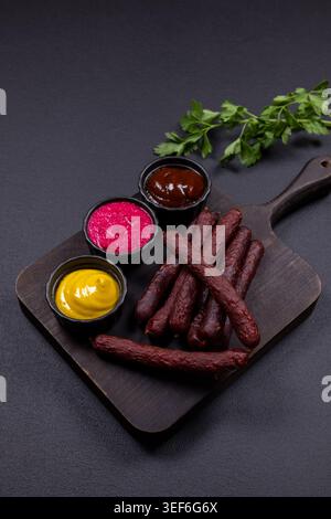 Geräucherte Würstchen auf einem dunklen Holzschneidebrett, serviert mit gelben Senfschüsseln, Rote-Rüben-Dip und Grillsoße, garniert mit frischer Petersilie Stockfoto
