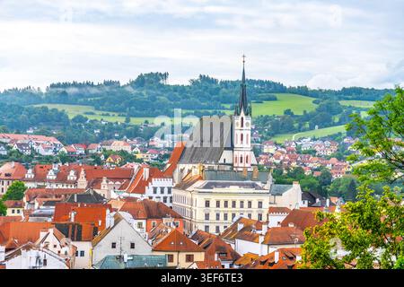 Die St.-Veit-Kirche steht hoch in Cesky Krumlov. Die Stadt verfügt über farbenfrohe Dächer und grüne Hügel im Hintergrund. Es ist ein Sommertag und die Gegend ist lebhaft mit Besuchern. Stockfoto