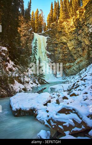 Gefrorener Wasserfall und Fluss in verschneiten Winterlandschaften mit Klippen und immergrünen Bäumen Stockfoto