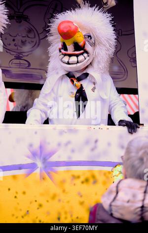 Fastnachtsfigur mit grinsender Larve, die Konfetti von einem Wagen wirft, gesehen von der Straße aus während des Cortège während der Basler Fasnacht 2026, Basel, Schweiz Stockfoto