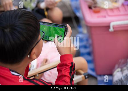 PHAYAO, THAILAND, 11. April 2026: Junge sitzt und spielt Spiel auf dem Smartphone, zwanglose Nutzung des Telefons im Freien, entspannter digitaler Moment, Nahaufnahme, Chiang Kham. Stockfoto