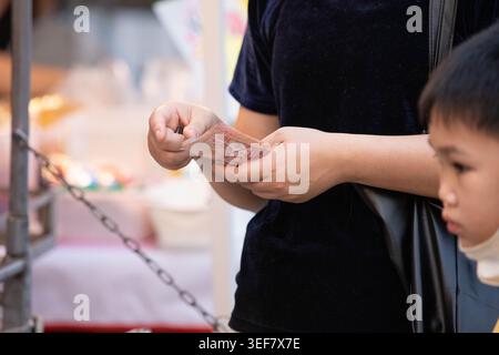 PHAYAO, THAILAND, 11. April 2026: Weibliche Hand, die Banknoten hält, um Waren auf dem Markt zu bezahlen, Menschen, die auf dem Freiluftmarkt einkaufen, Nahaufnahme, Chiang Kham. Stockfoto
