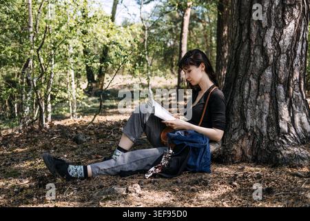 Die Person sitzt auf dem Boden, während sie ein Buch unter einem Baum im Wald liest. Digitale Entgiftung, langsame Lebensbewegung, Entkommen aus der Stadt, einfache Freuden. Stockfoto