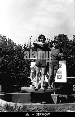 Sloviansk, 1983, Pionierlager Wishnegorsk in Slavkurort. Zwei Lagerberater posieren spielerisch barfuß auf einem Steinbrunnen. Der junge Mann beugt scherzhaft seinen Bizeps und zeigt seine Stärke, während die Frau neben ihm steht und einen Fußball hält. Beide tragen dazu passende sportliche Freizeitkleidung zu Shorts und T-Shirts. Die Szene spielt draußen vor üppigen Parkblattpflanzen und einem sowjetischen Poster. Dieses lebendige Bild zeigt den energischen und humorvollen Lebensstil des Jugendpersonals während der Sommerferien und fängt das Leben im friedlichen Donbass ein. Stockfoto