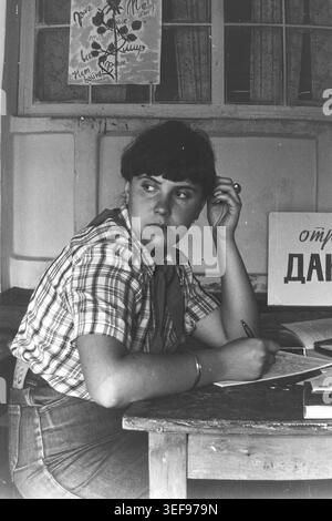 Sloviansk, 1983, Pionierlager Wishnegorsk in Slavkurort. Eine junge Beraterin sitzt drinnen an einem Schreibtisch und hält ihr Schreiben an, um mit einem nachdenklichen Ausdruck über ihre Schulter zu schauen. Sie trägt ein lässiges kariertes Hemd mit einer Pionierkrawatte. Ein Schild für „Squad DANKO“ und ein handgefertigtes Poster sind im Hintergrund zu sehen. Dieses ehrliche, ausdrucksstarke Porträt fängt die authentische Atmosphäre und die administrativen Aufgaben des Lagerpersonals während der sowjetischen Sommerferien ein und dokumentiert das Leben im friedlichen Donbass. Stockfoto
