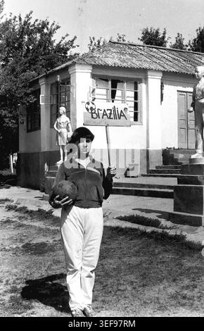 Sloviansk, 1983, Pionierlager Wishnegorsk in Slavkurort. Eine junge Beraterin posiert selbstbewusst vor einem Lagergebäude, hält ein handgefertigtes „BRAZILIA“-Schild und einen Fußball in der Hand. Sie trägt eine Mütze, eine Sportjacke und eine leichte Jogginghose. Zwei silberne Pionierstatuen flankieren den Eingang hinter ihr. Dieses stolze und sportliche Porträt fängt den Fokus des Camps auf Sportunterricht, kreative Spiele und den energiegeladenen Lebensstil des Jugendpersonals während der Sommerferien ein und zeigt das Leben im friedlichen Donbass. Stockfoto