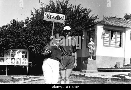Sloviansk, 1983, Pionierlager Wishnegorsk in Slavkurort. Zwei fröhliche junge Beraterinnen posieren mit einem handgefertigten hölzernen „BRAZILIA“-Schild und einem Fußball vor einem Lagergebäude. Sie stehen eng zusammen, tragen bequeme Sommersportbekleidung und unterstreichen ihre Freundschaft und ihren sportlichen Geist. Eine silberne Pionierstatue ist auf den Stufen des Gebäudes im Hintergrund zu sehen. Das Foto fängt perfekt den informellen Spaß, die Kreativität und die fröhliche Jugendkultur des friedlichen Donbas ein. Stockfoto