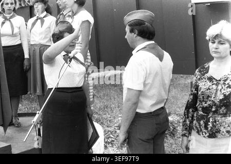 Sloviansk, 1983, Pionierlager Wishnegorsk in Slavkurort. Während der Eröffnungszeremonie des Camps steht eine Beraterin vor einem Mikrofon und hält eine Rede, während sie einen traditionellen Pioniersalut gibt. Neben ihr stehen ein Mann in Krawatte und eine andere Frau. Ein Pionier steht im Vordergrund. Die feierliche Veranstaltung findet in der Nähe eines großen architektonischen Bauwerks im Park statt. Dieses Foto zeigt die formale Hierarchie, Disziplin und patriotische Rituale der sowjetischen Jugendsommerlager und zeigt das Leben im friedlichen Donbass. Stockfoto