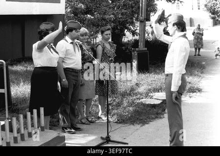 Sloviansk, 1983, Pionierlager Wishnegorsk in Slavkurort. Eine offizielle Versammlung zur Eröffnung der Sommerschicht findet im Freien statt. Ein junger Mann steht auf dem Spiel und überbringt den weiblichen Lagerführern, die sich um ein Mikrofon versammelt haben, einen formellen Pioniergruß. Das Personal trägt weiße Hemden, rote Krawatten und Uniformmützen. Die Szene vor einer Parkkulisse zeigt das strenge Protokoll, die Zeremonienkultur und die strukturierte Hierarchie, die typisch für sowjetische Jugendorganisationen ist, und dokumentiert einen authentischen Moment im friedlichen Donbass. Stockfoto
