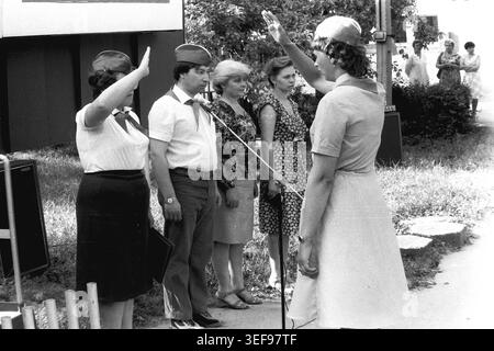 Sloviansk, 1983, Pionierlager Wishnegorsk in Slavkurort. Während der offiziellen Eröffnungszeremonie der Sommerschicht überbringt eine Lagerberaterin in einem Kleid dem am Mikrofon stehenden Lagerleitungsteam einen formellen Pioniergruß. Die Führer tragen traditionelle Uniformmützen und rote Krawatten und stehen förmlich für den Empfang des Berichts. Dieses Foto im Freien zeigt die strenge Disziplin, die ideologischen Traditionen und die strukturierte Organisation der sowjetischen Jugendlager, die das Leben im friedlichen Donbass zeigen. Stockfoto