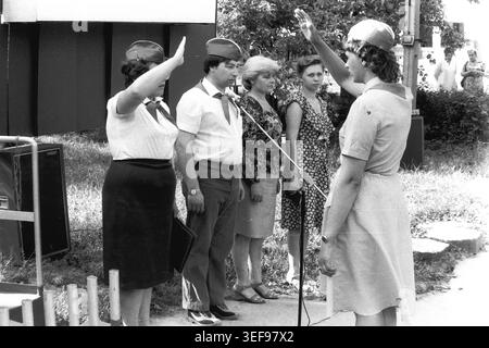 Sloviansk, 1983, Pionierlager Wishnegorsk in Slavkurort. Eine formelle Außenversammlung markiert die Eröffnung der Sommerschicht. Eine Lagerberaterin gibt einen Bericht ab, während sie dem Führungsteam am Mikrofon, das feierlich zurückgrüßt, einen Pioniergruß gibt. Sie tragen klassische sowjetische Sommeruniformen mit roten Krawatten und Kappen. Dieses dokumentarische Bild betont die zeremoniellen Protokolle, die starke Disziplin und den kollektiven Geist historischer Jugendorganisationen und fängt das Leben im friedlichen Donbass ein. Stockfoto