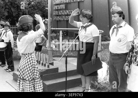Sloviansk, 1983, Pionierlager Wishnegorsk in Slavkurort. Die Lagerleiterinnen stehen an einem Mikrofon und sprechen während der Eröffnungszeremonie der Sommerschicht förmliche Pioniergrüße aus. Dahinter steht ein großes ideologisches Denkmal mit einem patriotischen Slogan über die Kommunistische Partei. Das Personal trägt traditionelle Uniformen mit roten Krawatten. Dieses eindrucksvolle Foto verkörpert perfekt die politische Bildung, die starre Struktur und die zeremonielle Atmosphäre der sowjetischen Jugendlager und zeigt den friedlichen Donbass. Stockfoto