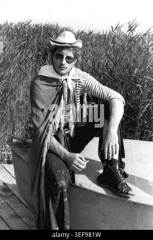 Sloviansk, 1983, Pionierlager Wishnegorsk in Slavkurort. Ein junger männlicher Lagerberater, kreativ gekleidet für das Neptun's Day Festival, posiert selbstbewusst, während er auf einem Boot inmitten von hohem Schilf sitzt. Er trägt ein gestreiftes Matrosenhemd, einen dunklen fransenumhang, eine Sonnenbrille und einen Cowboyhut, der eine Zigarette in der Hand hält. Dieses stilvolle und rebellische Dokumentarporträt fängt die ungezwungene Sommerfreizeit, den Theaterspaß und die Vintage-Jugendmode der 1980er Jahre ein und bewahrt das Leben im friedlichen Donbass. Stockfoto