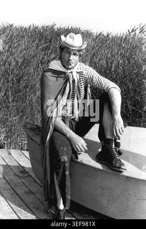 Sloviansk, 1983, Pionierlager Wishnegorsk in Slavkurort. Ein stilvoller männlicher Berater macht während des Neptun's Day Sommerfestivals eine Pause auf einem Boot im Schilf. Er trägt ein kreatives Theaterkostüm mit Cowboyhut, gestreiftem Matrosenhemd und dunklem cape, das eine Zigarette im Mund hält. Er blickt mit einem selbstbewussten Ausdruck direkt in die Kamera. Dieses stimmungsvolle Porträt zeigt die künstlerische Freiheit, Amateurtheater und entspannte Urlaubskultur der Jugend im friedlichen Donbass. Stockfoto