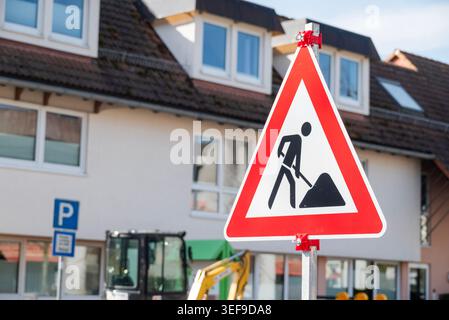 Nahaufnahme eines dreieckigen rot-weißen Straßenbauwarnschildes auf einer Stadtstraße. Im verschwommenen Hintergrund, ein Wohngebäude und ein gelbes Stockfoto