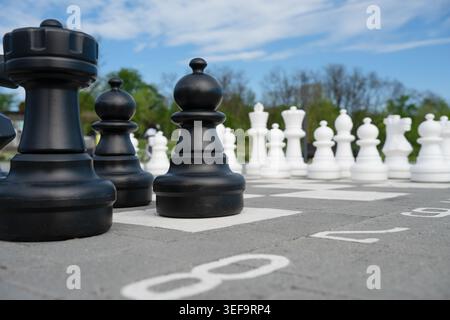 Niedriger Blickwinkel auf riesige schwarze Schachfiguren auf einem großen Outdoor-Schachbrett mit weißen Schachfiguren im Hintergrund unter blauem Himmel. Stockfoto