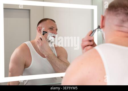 Hübscher junger Mann rasiert sich sein Gesicht mit Rasierer im Badezimmerspiegel während der Morgenroutine. Männerpflege, Hautpflege und Körperpflegekonzept. Stockfoto