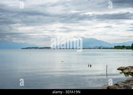 Ruhiges Seewasser mit fernen Städten und Bergen unter dramatischem bewölktem Himmel, friedliche natürliche Landschaft mit sanften Reflexen und gedämpften Tönen Stockfoto