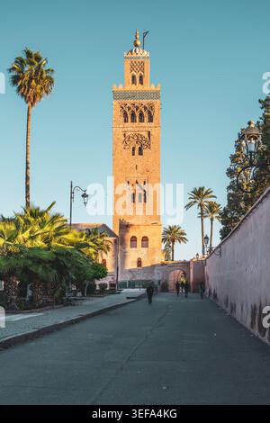 Die Menschen laufen entlang des Weges in der Nähe der Kutubiyya Moschee in Marrakesch. Die Sonne untergeht hinter der Moschee und wirft ein warmes Licht auf die Gegend. Palmen säumen die Straße und tragen zur Atmosphäre bei. Stockfoto