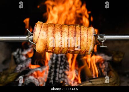 Porchetta wird auf Rotisserie über offenem Holzfeuer mit knusprigem Knistern und Flammen gebraten. Traditionelles langsam gekochtes Schweinebraten, rustikales Gourmet-Fleischgericht Stockfoto