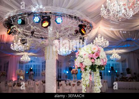 Eleganter Veranstaltungsort, dekoriert für Hochzeit oder Gala. Rosafarbenes und weißes Blumenmuster im Vordergrund. Vorhangdecke mit Kristallleuchtern und Farbtönen Stockfoto