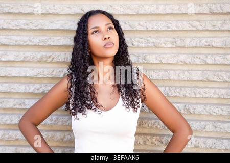 Die lebendige junge Frau mit lockigen Locken steht in Sommerkleidung selbstbewusst vor strukturiertem Hintergrund Stockfoto