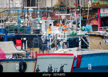 Yilan, Taiwan - 31. März 2025: Fischer reparieren Netze an Deck im Hafen von Nanfangao, mit vertäuten Schiffen und einem taiwanesischen Tempel im Hintergrund. Stockfoto
