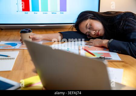 Erschöpfte Büroangestellte, die auf ihrem Schreibtisch schlief, nachdem sie spät in der Nacht in einem schwach beleuchteten, modernen Arbeitsplatz gearbeitet hatte. Stockfoto