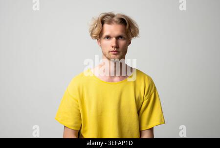 Junger blonder Mann mit ernstem Ausdruck, der direkt in die Kamera schaut, lässiges gelbes T-Shirt, Studio-Porträt auf neutralem grauen Hintergrund Stockfoto