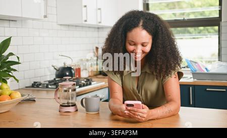 Lächelnde junge Frau in der Küche mit Smartphone und Kaffee auf der Arbeitsplatte während der Morgenroutine Stockfoto